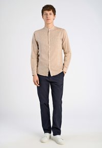 Camicia a maniche lunghe color marrone chiaro con bottoni e tasca, abbinata a pantaloni blu scuro e sneakers bianche, presentata su uno sfondo neutro.
