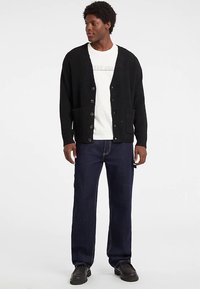 Čierny pletený kardigán s výstrihom do V, prednými gombíkmi a vreckami, nosený cez bielu košeľu a kombinovaný s džínsami a čiernymi topánkami.