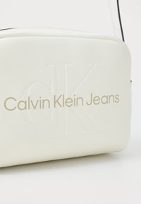 Borsa in ecopelle bianca con logo "Calvin Klein Jeans" in rilievo dorato. Texture liscia; forma rettangolare; tracolla regolabile.