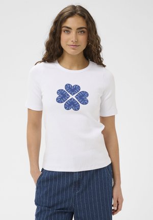 Ženska, oblečena v belo majico z kratkimi rokavi, okrašeno z modrim abstraktnim srčkastim cvetličnim vzorcem, in modre hlače z ozkimi črtami, stoji z rokami v žepih.