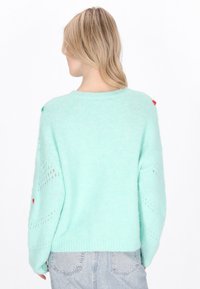 Maglione in maglina verde menta, morbido e dalla vestibilità rilassata, con disegni traforati sulle maniche e piccoli accenti colorati sulle spalle.