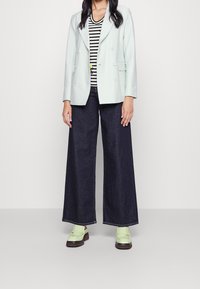 Blazer azul claro de doble botonadura sobre una camisa a rayas blanca y negra, combinado con pantalones de mezclilla oscuros de pierna ancha y mocasines verdes.