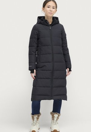 Sort lang pufferjakke med horisontale syninger, lynlås foran og hætte. Båret over blå jeans og hvidt fodtøj med pelsdetaljer.