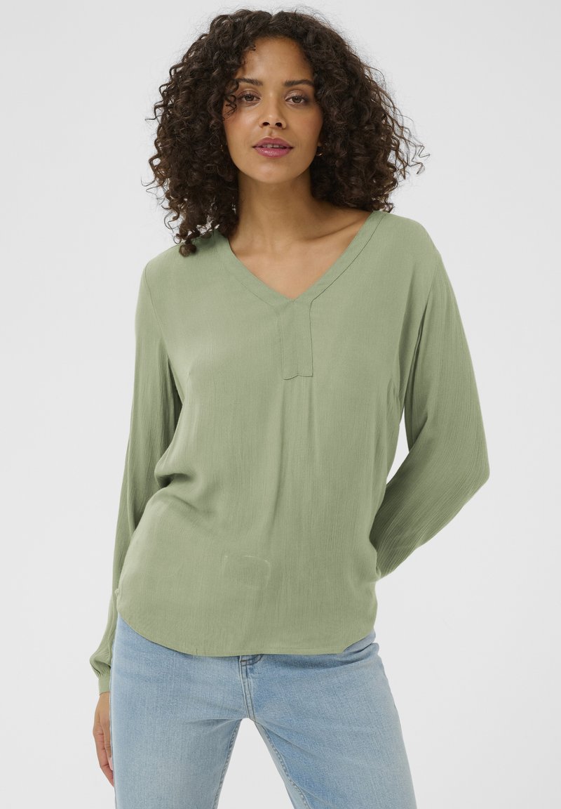 Blouse à manches longues et col en V en tissu vert clair, présentant une finition légèrement texturée et une coupe décontractée, associée à un jean bleu.