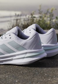 Paire de chaussures de course blanches Adidas avec des rayures vert clair et des semelles Cloudfoam reposant sur un trottoir à l'extérieur près de plantes floues.