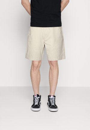 Homme portant un short beige jusqu'aux genoux, des baskets montantes noires à lacets blancs, et une chemise noire, debout contre un mur uni.