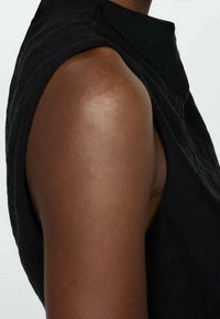 Zwarte mouwloze top met een hoge kraag en textuurstof, die de schouder- en bovenarmzone accentueert tegen een effen achtergrond.