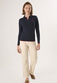 Marineblauwe polo met lange mouwen en een kraag, met een klein rood hart-embleem op de borst en contrasterende rode bies aan de manchetten.