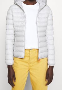 Doudoune gris clair avec capuche, présentant des coutures horizontales, une fermeture éclair et des accents orange, associée à un jean jaune.