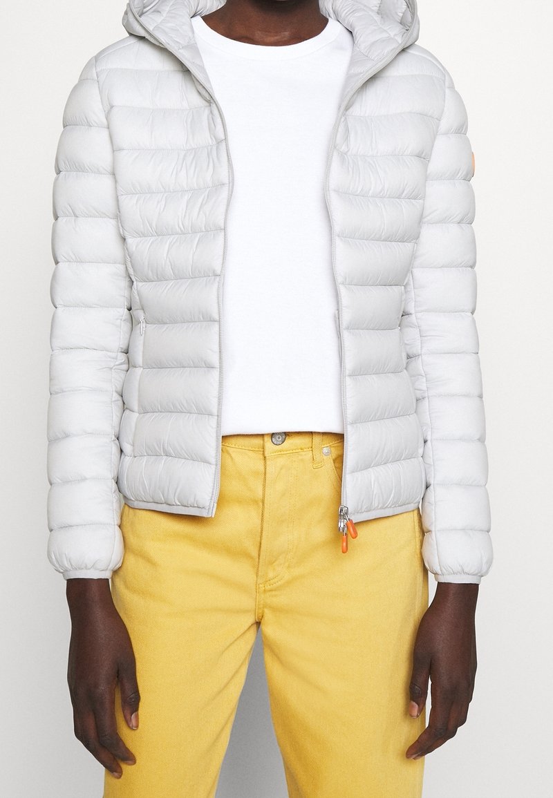 Doudoune gris clair avec capuche, présentant des coutures horizontales, une fermeture éclair et des accents orange, associée à un jean jaune.