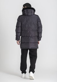 Padded czarny płaszcz z kapturem wyłożonym futrem, z delikatnym wzorem logo, w oversize'owym fasonie i długimi rękawami. Nosi się go z czarnymi spodniami i białymi butami.