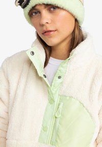 Light green and cream fleece jacket with a zippered pocket and snap closures. Features a soft texture and oversized collar.