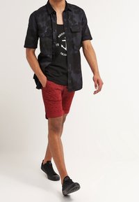 Mężczyzna idący, ubrany w czarną koszulę z krótkim rękawem na guziki, na czarnym podkoszulku, czerwone szorty w wzory oraz czarne tenisówki, na białym tle.