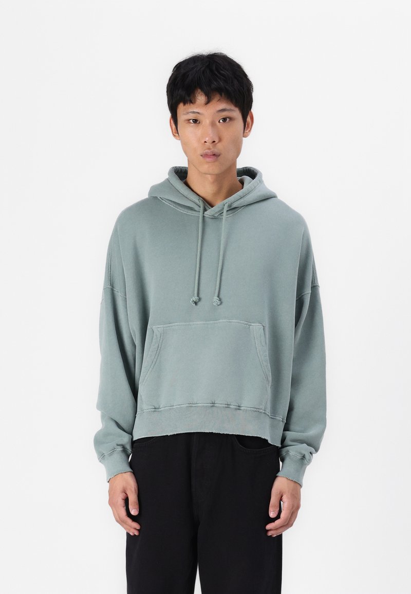 Lysgrøn cropped hoodie lavet af blødt stof, med en frontlomme, snoretræk ved hætten og ribbede manchetter. Bæres med sorte bukser.