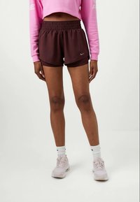 Vrouw in bordeauxrode Nike hardloopshorts, witte Nike sokken, lichtroze Nike sneakers en een kort gesneden roze lange mouw shirt, staande tegen een plain witte achtergrond.