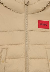 Beige Daunenjacke mit vorderem Reißverschluss und zwei Druckknopf-Taschen, mit einem roten Aufnäher mit "HUGO" in schwarzem Text auf der Brust.