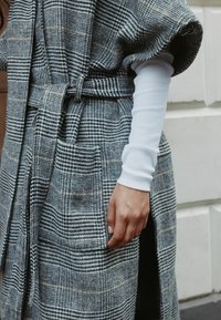 Hall hallikontrolliga vööga mantel, mis on valmistatud tekstuurse kangaga, küljetaskutega ning mille avatud varrukast paistab valge pika varrukaga särk.