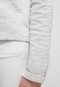 Persona che indossa una maglietta a maniche lunghe grigia chiaro con i polsini arrotolati sopra dei pantaloni bianchi, con il braccio rilassato lungo il fianco.