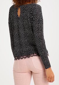 Kobieta ubrana w czarną bluzkę z długim rękawem w serduszka z koronkowym wykończeniem i jasnoróżowe dżinsy, pokazana od tyłu.