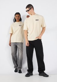 Beige t-skjorter med kort erm og svart trykk, grå løstsittende jeans og svarte bukser, båret med sporty svarte sko mot en hvit bakgrunn.