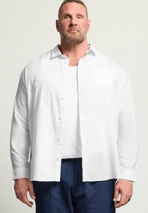 Brodaty mężczyzna w rozpiętej białej koszuli na białym t-shircie i ciemnoniebieskich spodniach, stojący na tle jednolitego jasnego tła.