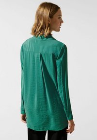 Long-sleeve green blouse featuring a buttoned collar, textured fabric, pleat detail at the back, and buttoned cuffs.