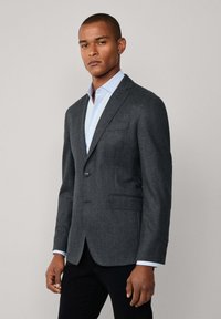 Charcoalgrauer Blazer aus strukturiertem Stoff, Ein-Knopf-Verschluss, zwei Fronttaschen und darunter ein kontrastierendes hellblaues Hemd.
