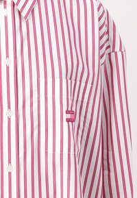 Vit skjorta med knappar och vertikala rosa ränder, en ficka på vänster bröst och en liten röd "TOMMY JEANS"-logotyp. Slät tygtextur.