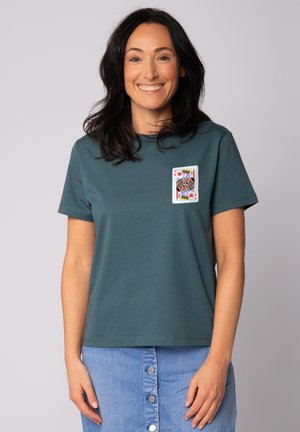 Lachende vrouw met donker haar die een turquoise T-shirt draagt met een afbeelding van de hartenkoning speelkaart en een lichtblauwe spijkerrok met knopen.