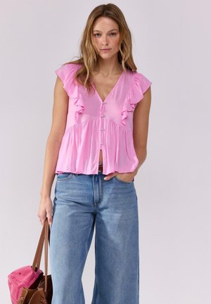 Blusa rosa con maniche corte a volant, scollatura a V e chiusura con bottoni, abbinata a jeans a gamba larga blu e una borsa marrone.