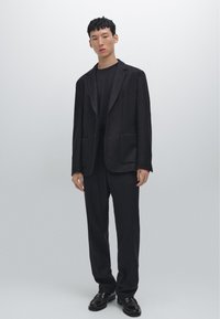 Blazer noir avec revers crantés, deux poches avant ; associé à un t-shirt noir et un pantalon ; le mannequin porte des chaussures noires ; texture lisse.