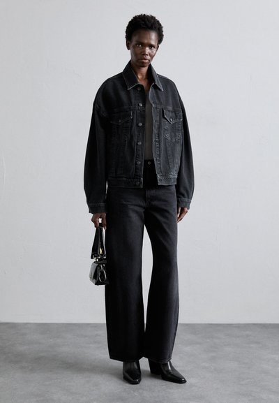 Black denim jacket with a relaxed fit, large front pockets, paired with wide-leg black jeans. Accessories include a small black handbag.