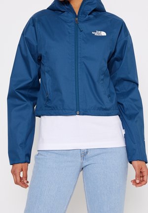Person iført en blå hættetrøje med lynlås fra The North Face, kombineret med en hvid skjorte og lyseblå jeans.
