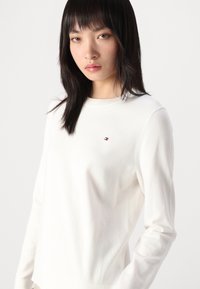 Maglione bianco lavorato a maglia con maniche lunghe, collo rotondo e un piccolo dettaglio del logo sul petto. Tessuto morbido e design aderente.