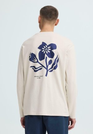 Man wearing beige long-sleeve shirt with large blue flower graphic and text "ARCHIVE of SOLID © 1999" on the back.