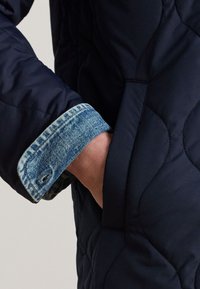 Hand in Tasche einer schwarzen gesteppte Jacke mit sichtbar blauem Jeanshemdbund darunter.