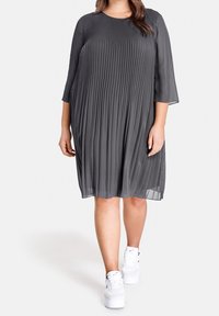 Vestido gris con plisados, mangas tres cuartos, escote redondo y textura fluida, combinado con zapatillas blancas para un look casual.