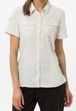 Femme portant une chemise beige clair à manches courtes avec deux poches poitrine et un pantalon noir, debout devant un fond uni.
