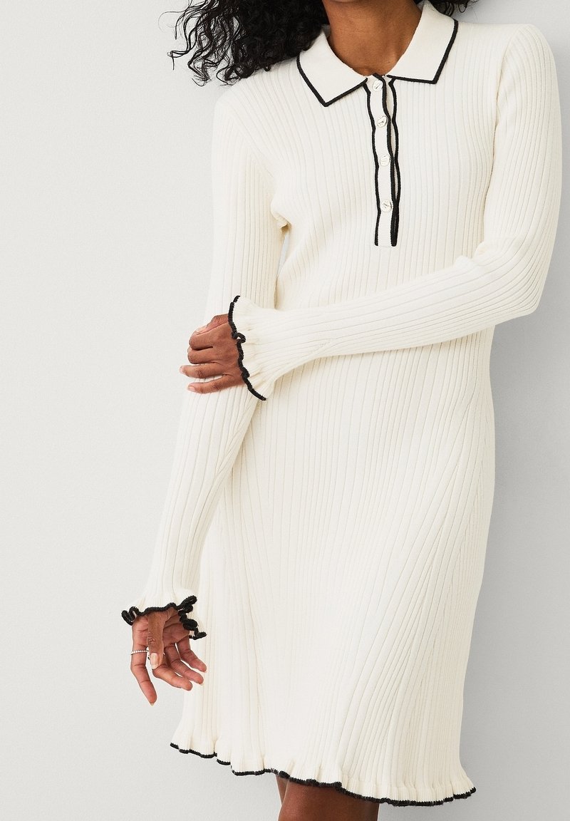 Femme portant une robe en maille côtelée blanche avec des bordures noires au col, à la patte de boutonnage, aux poignets et à l'ourlet, debout devant un fond uni.