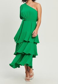 Zelené jedno-ramenné šaty s vrstvenými volány, vyrobené z texturované látky. Obsahují pásek v pase a mají délku po kotníky. Sandály jsou součástí.