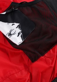 Tessuto leggero rosso con una sezione in rete nera texturizzata. Un'etichetta in stoffa decorata presenta un design di ragno nero su uno sfondo bianco.