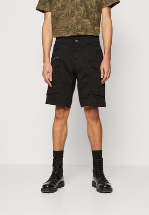 Homme portant un short cargo noir avec poches latérales, associé à des bottes noires lacées et une chemise à manches courtes à motifs vert olive.