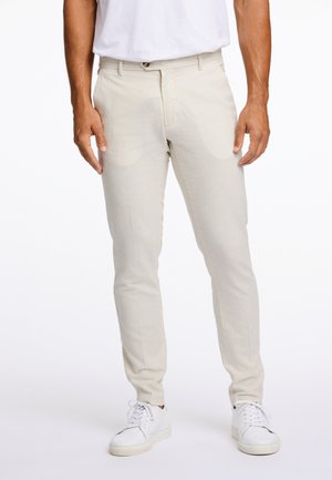 Pantalones beige, ligeros, con corte ajustado y frente plano. Cuentan con bolsillos laterales y cierre de botón. Llevados con zapatillas blancas.