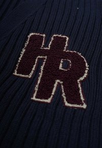Marinblått ribbat trikåtyg med en vinröd broderad "HR"-logotyp som är inramad i krämfärg. Texturerad yta med synliga stygn.