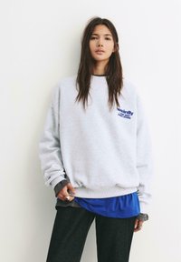 Felpa grigia con la scritta "weirdly", indossata sopra una maglietta in rete blu. Presenta una vestibilità ampia, polsini a coste e scollatura rilassata, abbinata a pantaloni scuri.