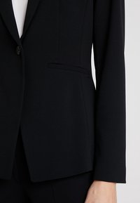 Blazer noir sur mesure avec un seul bouton et une poche passepoilée porté sur un haut blanc, montré sur le bras droit et la zone du torse.