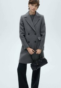 Double-breasted gray wool coat with large black buttons, notched lapels, and side pockets; paired with black pleated trousers and a black handbag.