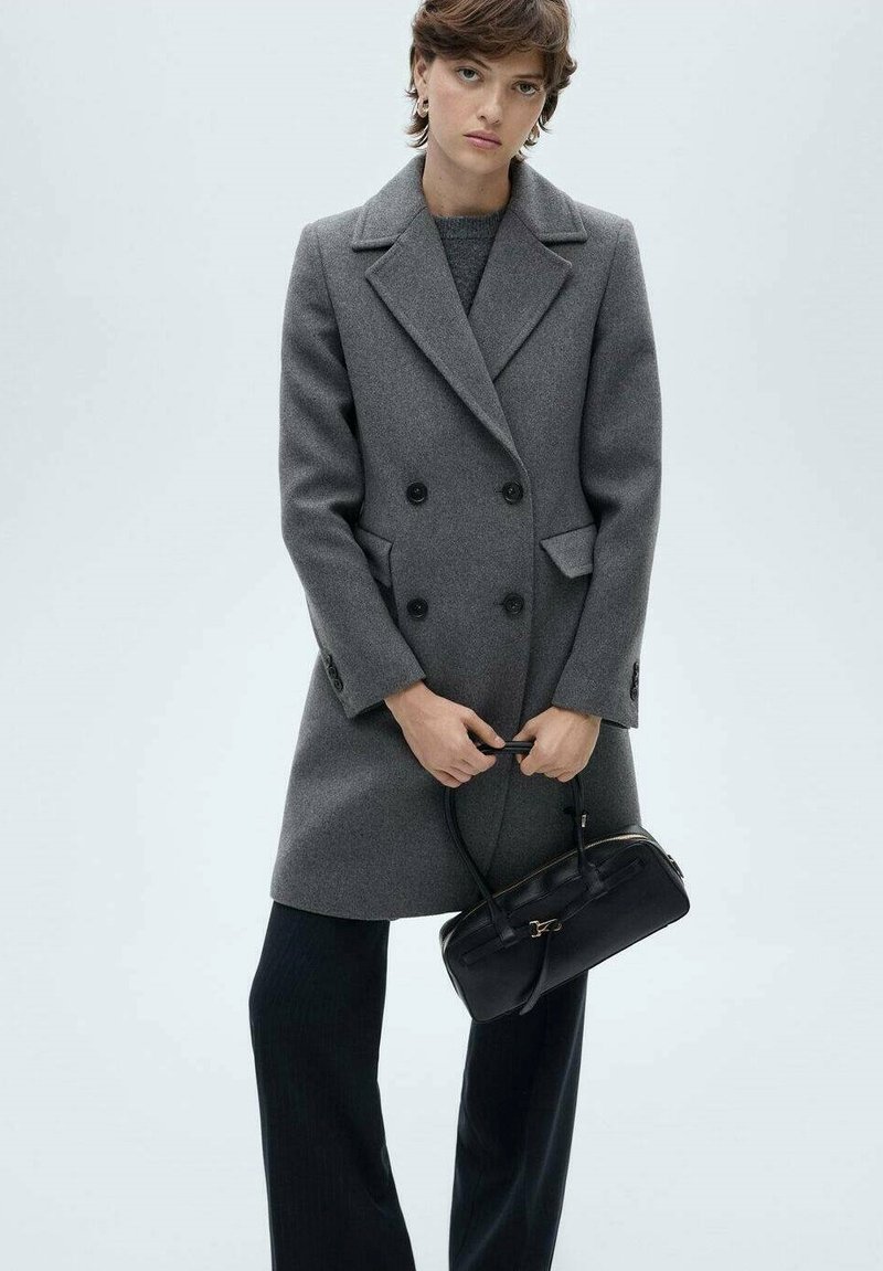 Double-breasted gray wool coat with large black buttons, notched lapels, and side pockets; paired with black pleated trousers and a black handbag.