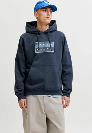 Homme portant un sweat à capuche bleu marine avec le logo "Jack & Jones", un pantalon beige et une casquette de baseball bleue, regardant sur le côté devant un fond uni.