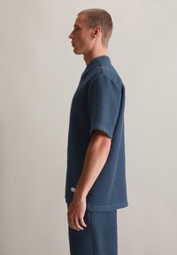 Blaues, kurzärmliges Hemd aus strukturiertem Stoff mit kragenlosem Ausschnitt und Knopfleiste. Getragen mit passenden Hosen, seitliche Ansicht gezeigt.
